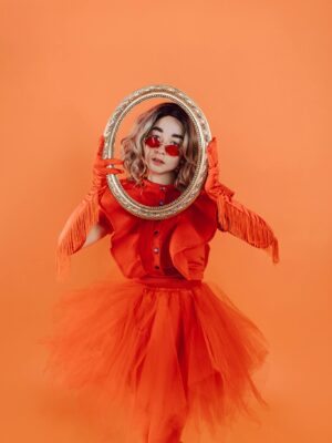 A person in an orange costume holding an ornate mirror against an orange background.