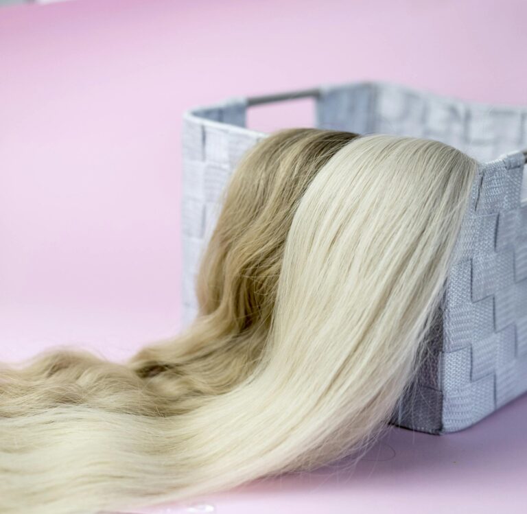 Close-up of blond and brown hair extensions draped over a woven basket, on a pink background.